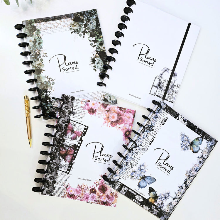 Four dream.design.bloom 2027 discbound planner notebooks with floral and vintage-style covers are arranged on a light surface with a gold pen nearby. The covers show the visible brand text 'Plans Sorted.