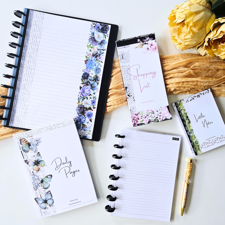 Flat-lay of stationery on a white surface: an open discbound notebook with lined pages, a closed notebook with a floral border reading 'Daily Pages', a shopping list pad, a 'Little Notes' card, a gold pen and dried yellow flowers.