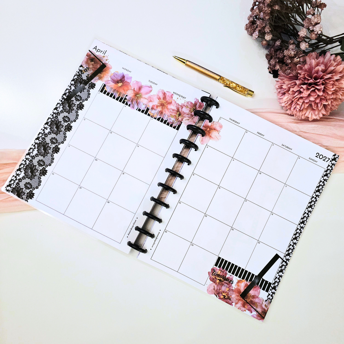 An open monthly planner spread on a white surface, decorated with pink floral graphics, black lace trim, and matte black discbound discs. A gold pen and dried flowers are placed nearby as styling props.