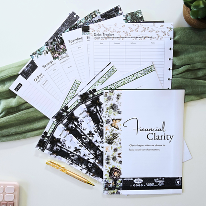 A styled flat-lay of a 'Financial Clarity' budgeting planner set on a white surface with green fabric and a small plant. The set includes a main notebook with floral and gold foil accents, several matching tracker sheets (Debt Tracker, Savings, Spending, Debt Paying, Debt Free Goals), a gold pen, and a small pink calculator. Disc Bound Planner. Discbound
