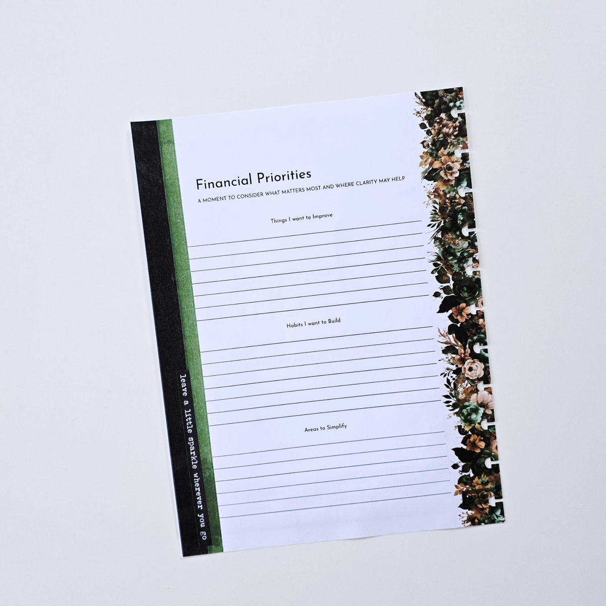 A flat-lay view of a 'Financial Priorities' notepad on a light gray background. The notepad has a white writing surface with lined sections, a black spine band with green and white text, and a floral border on the right side. Disc Bound Planner. Discbound