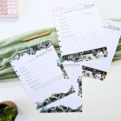 A styled flat-lay of several printed budget and savings goal tracker sheets with floral greenery borders, arranged on a white surface with a folded sage green cloth, a gold pen, a small plant, and part of a pink calculator visible. Disc Bound Planner. Discbound