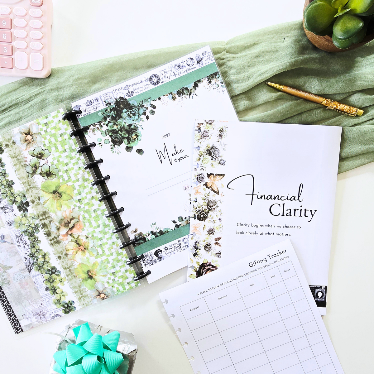 A styled flat-lay of a floral planner binder with a green and white patterned cover, open to a page that reads 'Make a plan.' A printed sheet titled 'Financial Clarity' and a 'Getting Tracker' page are placed beside it on a white surface with a folded sage-green fabric backdrop. Disc Bound Planner. Discbound