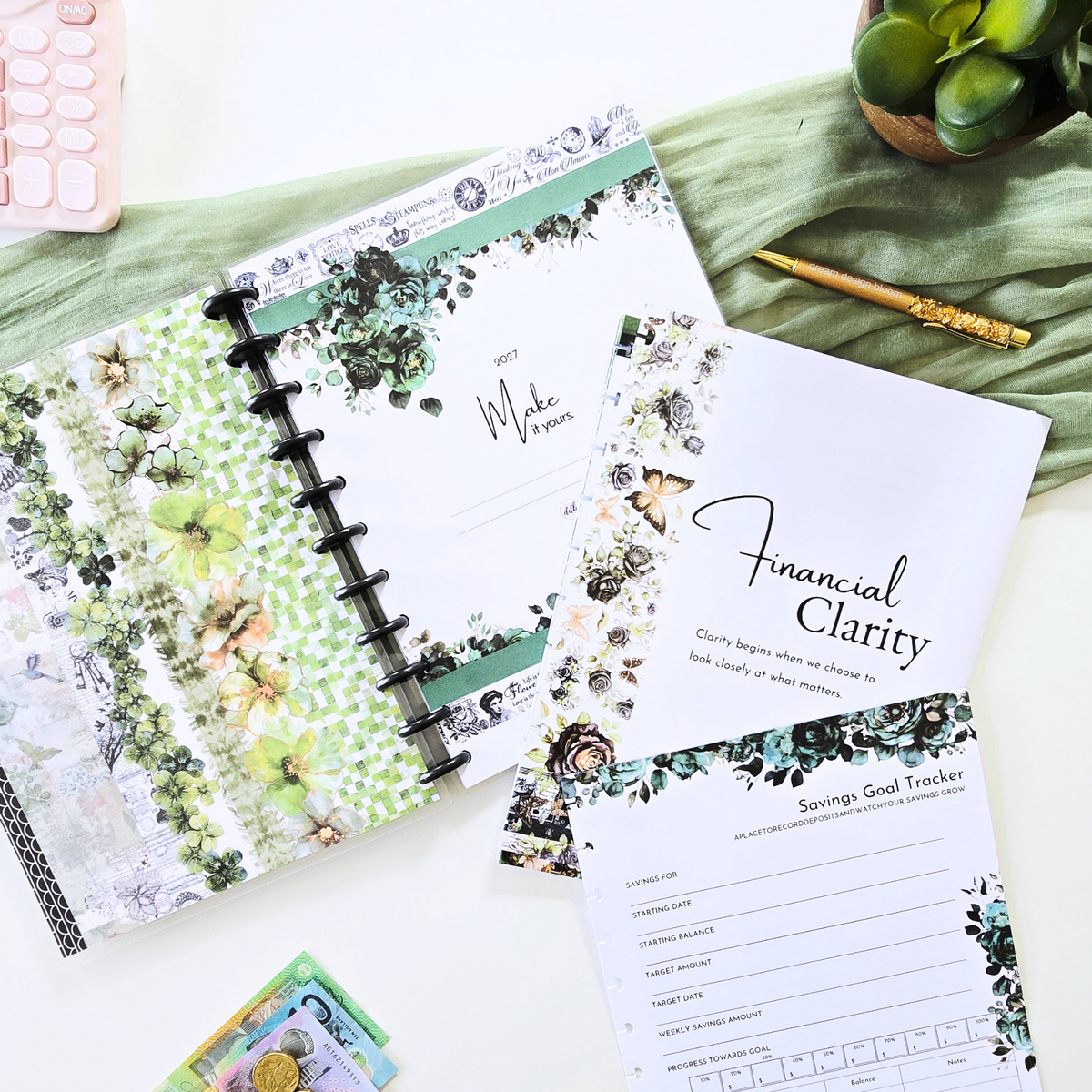 A styled flat-lay of floral planner inserts and budgeting sheets on a white surface with a green fabric backdrop. Visible items include an open ring-bound planner page with green and white botanical patterns, a sheet reading 'Financial Clarity,' a 'Saving Goal Tracker' sheet, a pink calculator, a gold pen, and a succulent. Disc Bound Planner. Discbound
