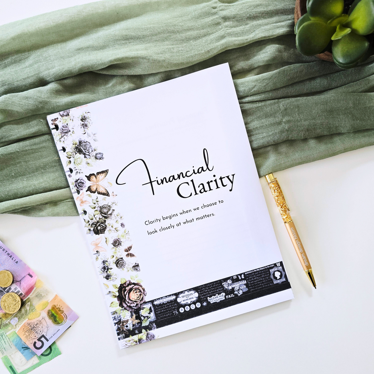 A styled flat-lay of a white notebook titled 'Financial Clarity' with floral illustrations on the left side, placed on a white surface with a sage green fabric backdrop, a gold pen, Australian coins, and a small succulent. Disc Bound Planner. Discbound