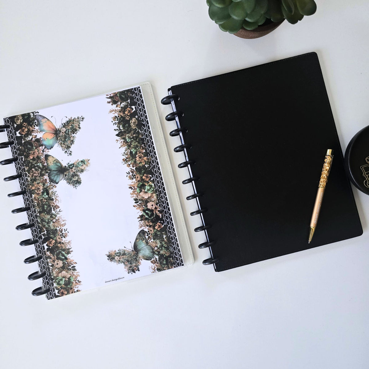 A black discbound planner or notebook lies on a light surface next to a floral planner cover with butterflies and the words. A gold crystal pen rests on the black discbound notebook or planner, with a small potted succulent and part of a black bowl visible in the scene. Disc Bound