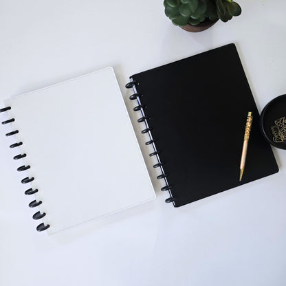 Two disc bound notebooks or planners on a white surface, one white with black spiral binding and one black with black spiral binding. A gold crystal pen and a small black dish with gold geometric designs rest on the black discbound notebook, with a small potted succulent in the top corner.
