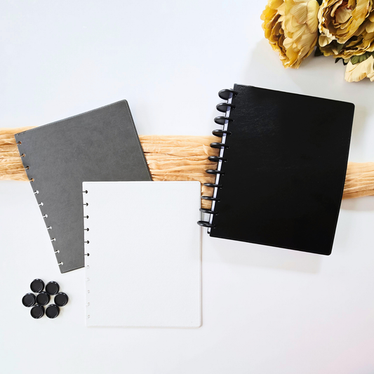 Three discbound notebooks in black, dark grey, and off-white are arranged on a light beige surface with a white rose and small succulent in the corners. The off-white notebook has gold text reading 'dream. design. bloom' near the bottom.