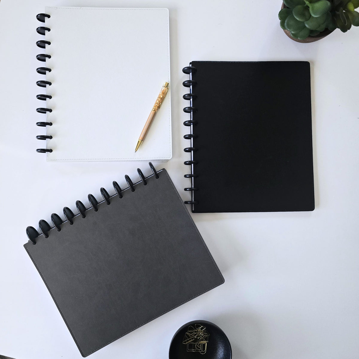 Three discbound leather covers in white, black, and dark gray are arranged on a white desk with a gold crystal pen, a small succulent, and a black dish.