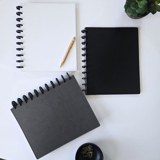 Three discbound leather covers in white, black, and dark gray are arranged on a white desk with a gold crystal pen, a small succulent, and a black dish.