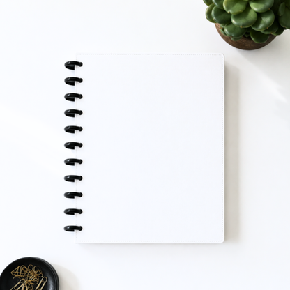 A white disc bound planner or notebook with black rings, photographed from above on a light neutral surface with a small potted succulent and paper clips nearby.
