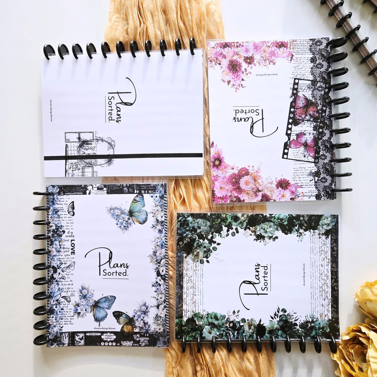 Four discbound planner notebooks with black metal spiral binding, arranged in a 2x2 grid on a light beige surface with decorative dried grass and gold flowers. The notebooks feature different floral and botanical designs on white backgrounds, with the text 'Plans Sorted' printed on each cover. Disc Bound Planner